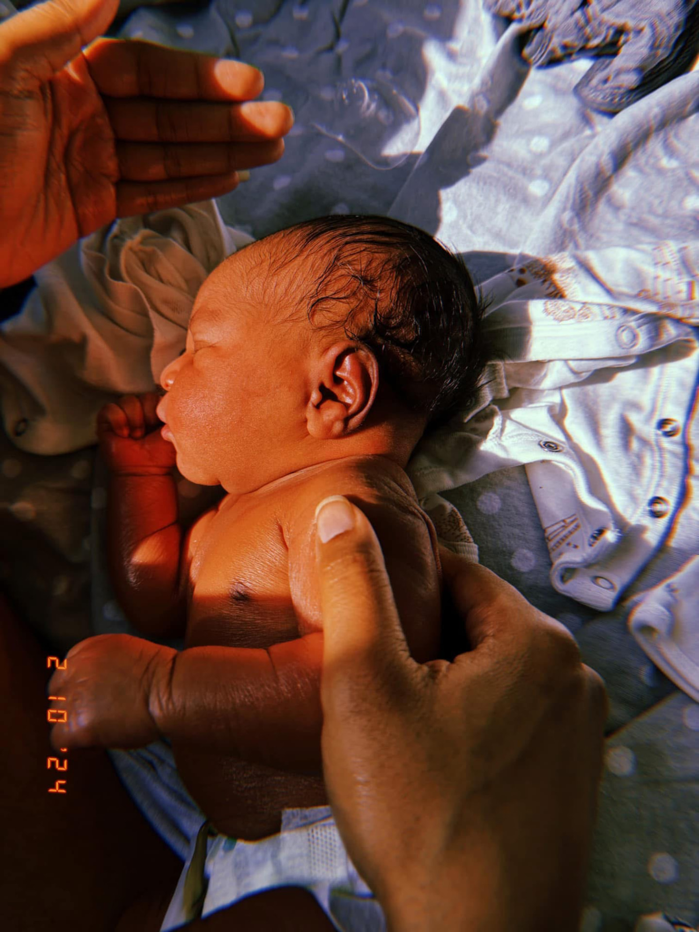 Newborn resting on soft bedding with an adult's hands gently supporting the baby