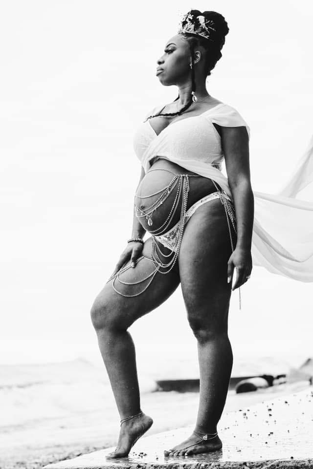 Omi standing barefoot by the water during pregnancy, her white dress flowing in the wind